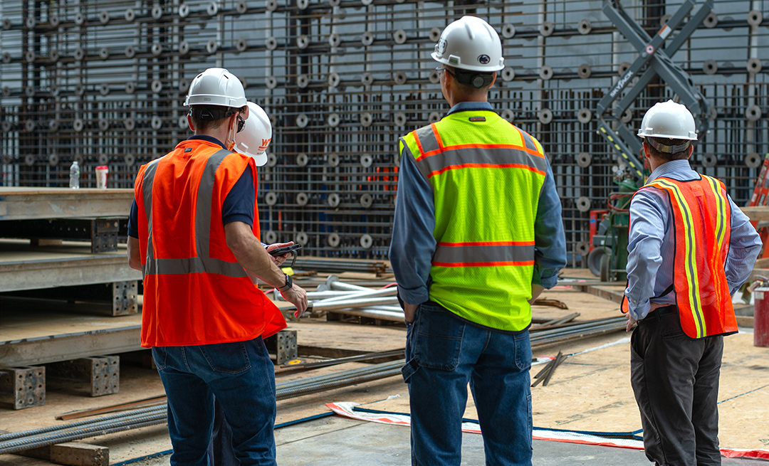 Four people in hard hats and neon safety vests converse while standing in a construction zone